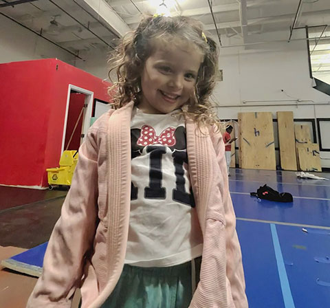 A smiling child in a pink cardigan and a shirt with a bow is standing indoors on a blue floor near a red structure.