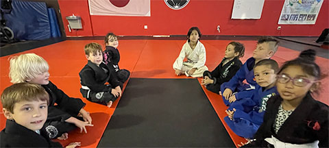 Children sit in a circle on red mats wearing martial arts uniforms, with flags displayed on the wall behind them.